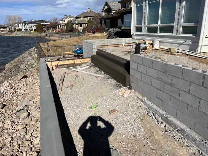 Construction site near a body of water.  Grey concrete block retaining wall and foundation being built next to a house.