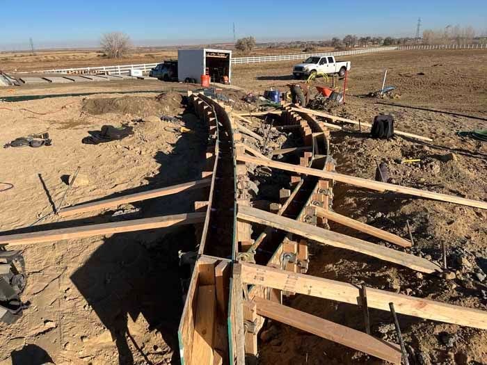 Construction site with curved concrete formwork.  A trench and wooden supports are visible. A truck and trailer are in the background.