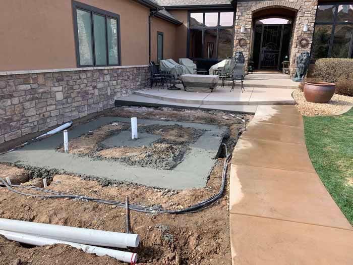Concrete foundation poured for a square feature with plumbing, alongside a house exterior and pathway.