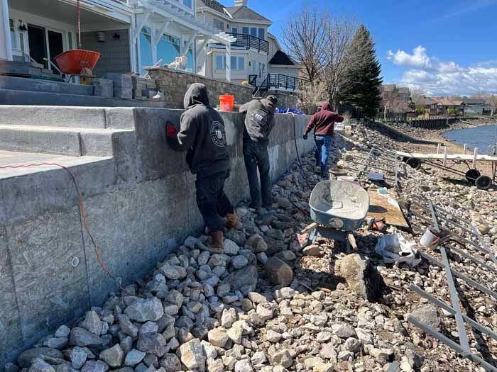 Three workers build a stone wall near water, next to a large house.