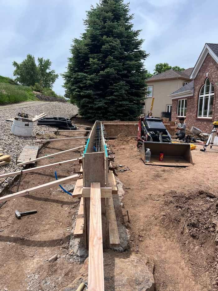 Construction site with wooden forms for a retaining wall, dirt trench, and equipment. A large evergreen tree and a house are in the background.