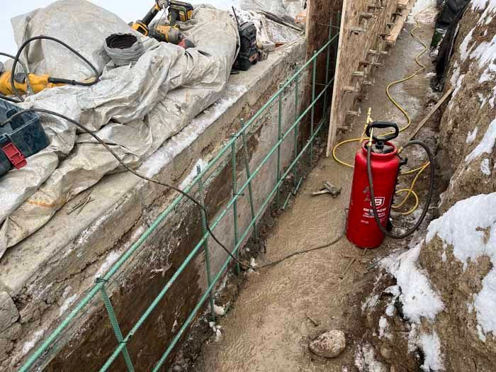 Construction site: rebar installed in trench, concrete forms, red sprayer, tools. Winter setting with snow.