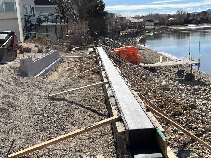 Construction site along a lake; concrete forms for a wall. Rebar and wooden supports are visible. Gray and brown tones.