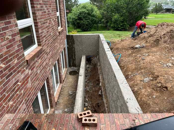 Exterior view of a brick house with concrete retaining wall construction, dirt excavation, and a person working.