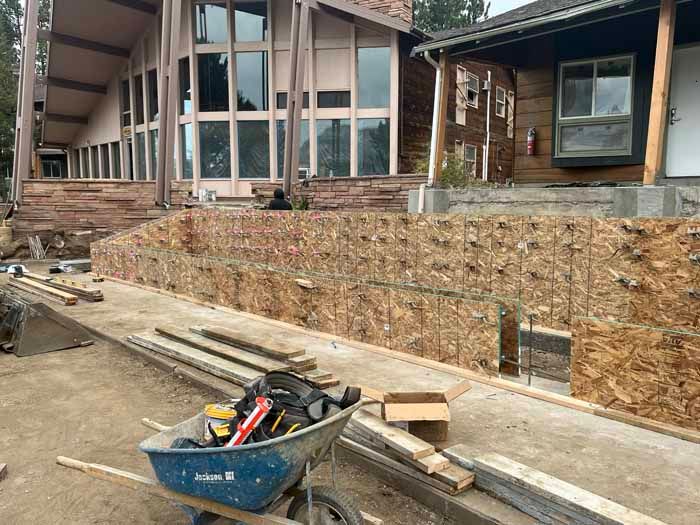 Construction site with plywood forms for concrete wall; house in background, wheelbarrow in foreground.