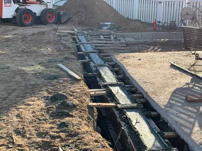 Trench for concrete work. Wooden forms are set, awaiting concrete. A Bobcat and pile of dirt are in the background.