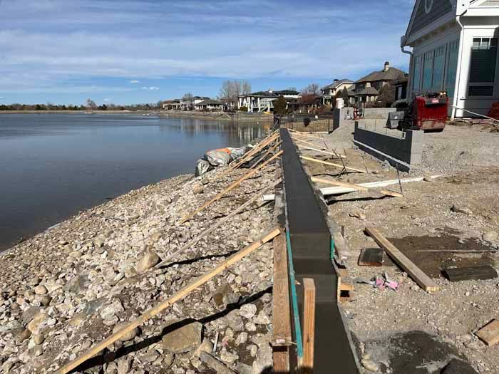 Lakeside concrete wall construction with wood supports.  Rocky shoreline, houses in background under blue sky.