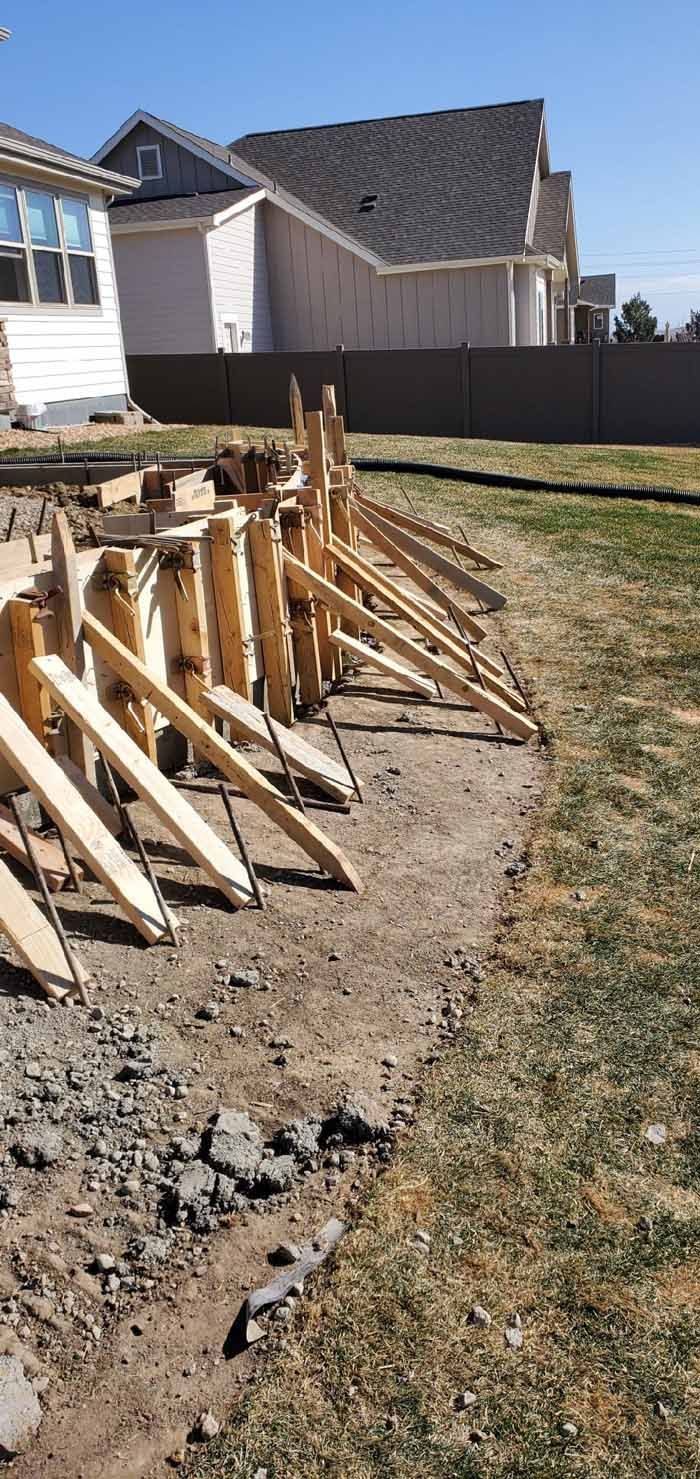 Wooden forms supported by angled braces, outlining a construction project in a yard with houses in the background.