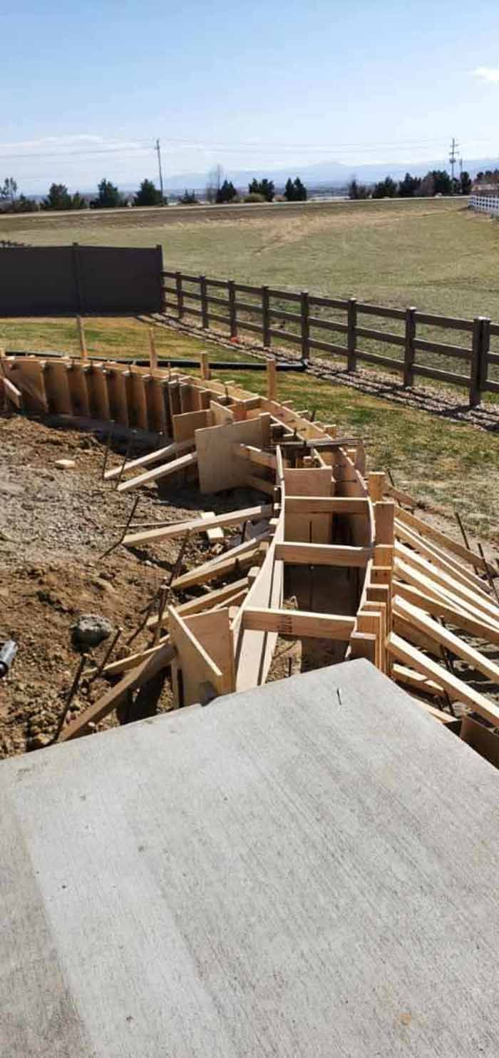 Construction site: Wooden forms for a concrete structure, next to a paved area and a grassy field with a fence.