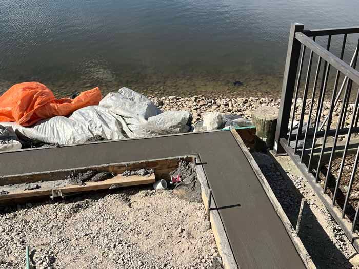 Newly poured concrete pathway next to water, with wood forms, rocks, and a metal railing.