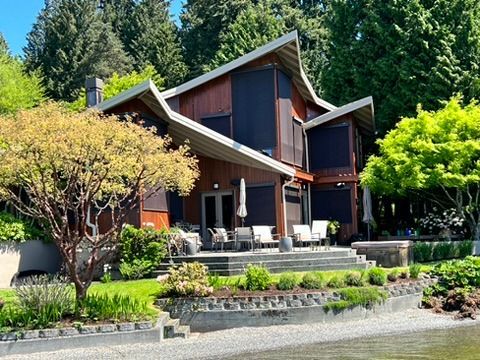 A multi-level wood-sided home with dark window shades and a patio, nestled among trees beside a pebble beach.