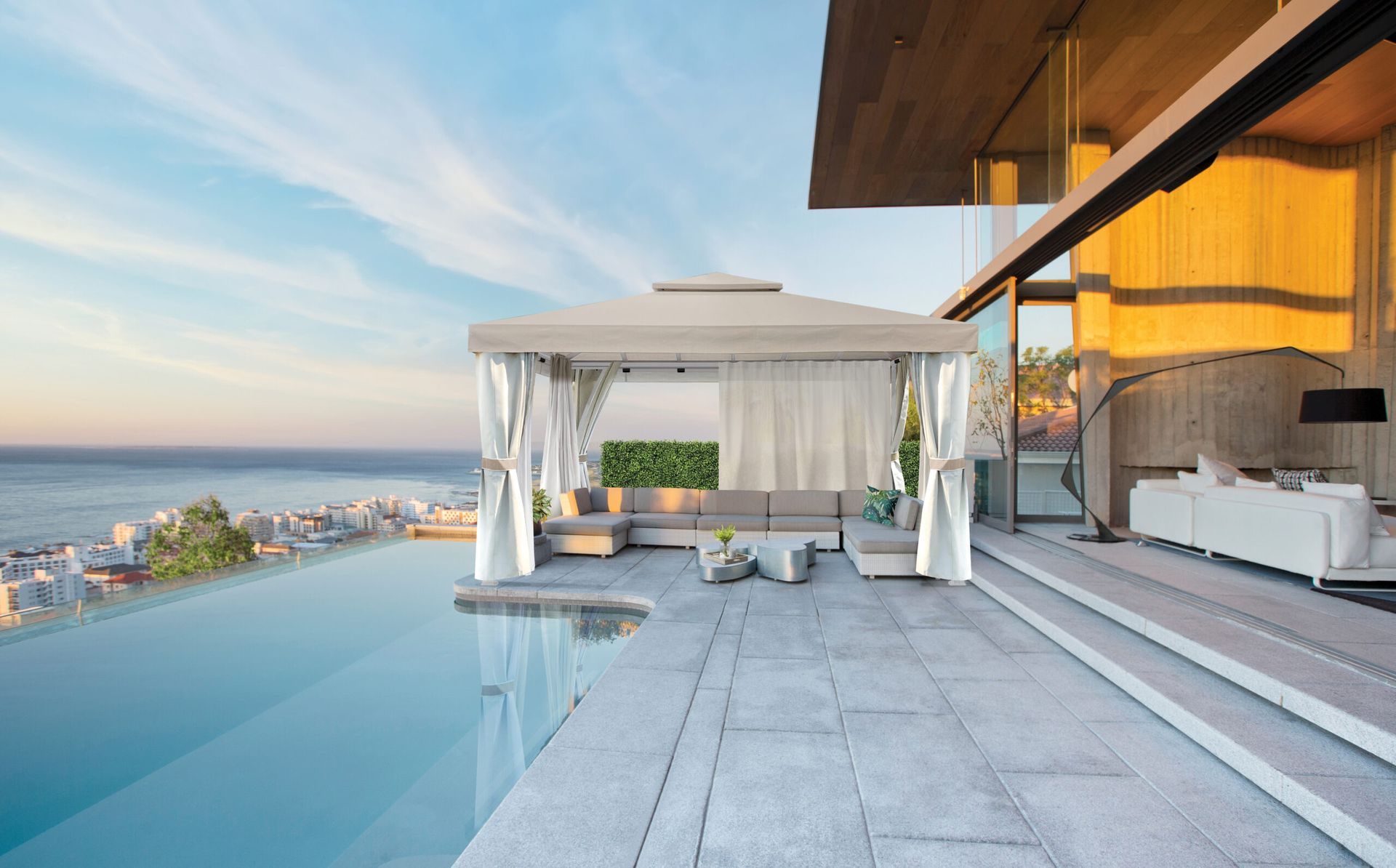 A beige gazebo with open curtains stands on a stone patio by an infinity pool overlooking a coastal city at sunset.