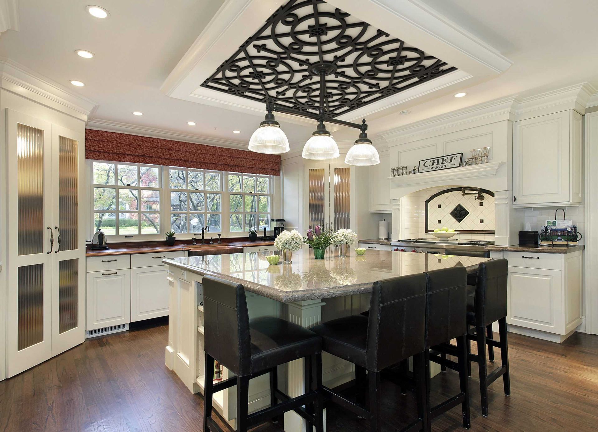 A white kitchen with a large granite-topped island, black bar stools, a decorative ceiling fixture, and wood flooring.