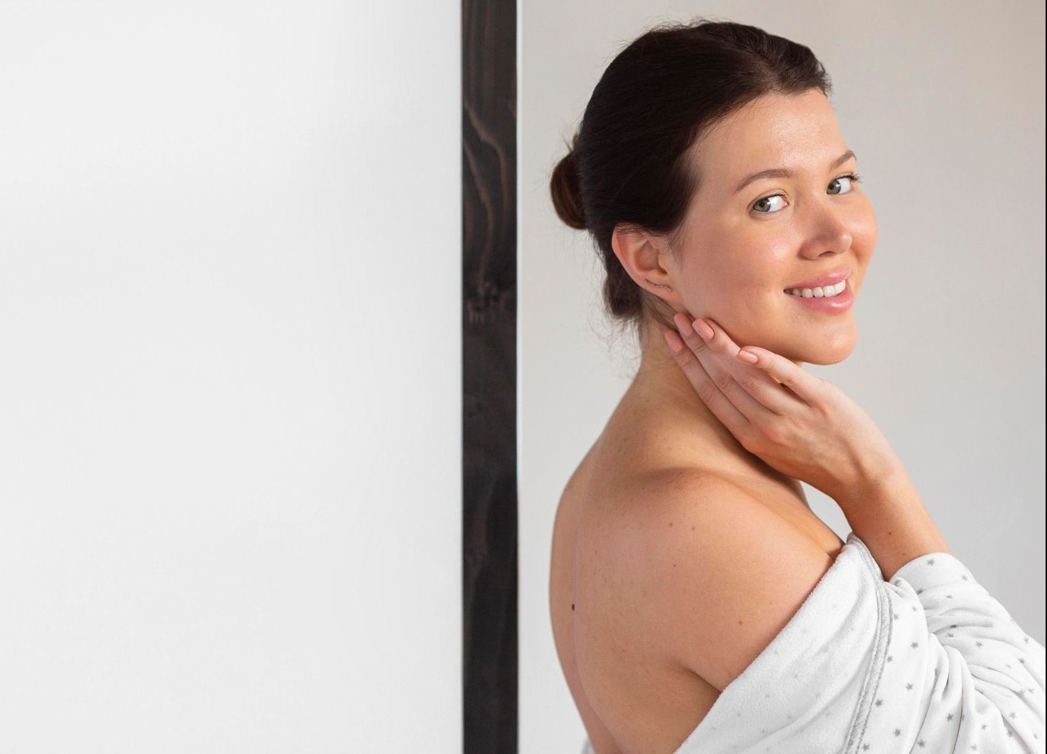 A woman in a white robe is smiling and touching her face