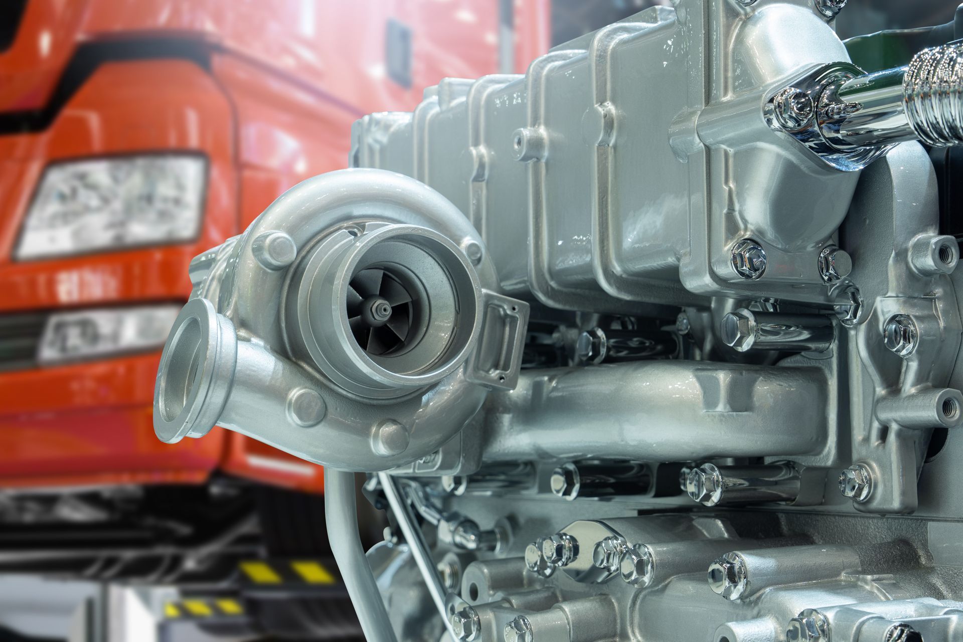 Close-up of a silver turbocharger attached to an engine, with an orange truck in the background.