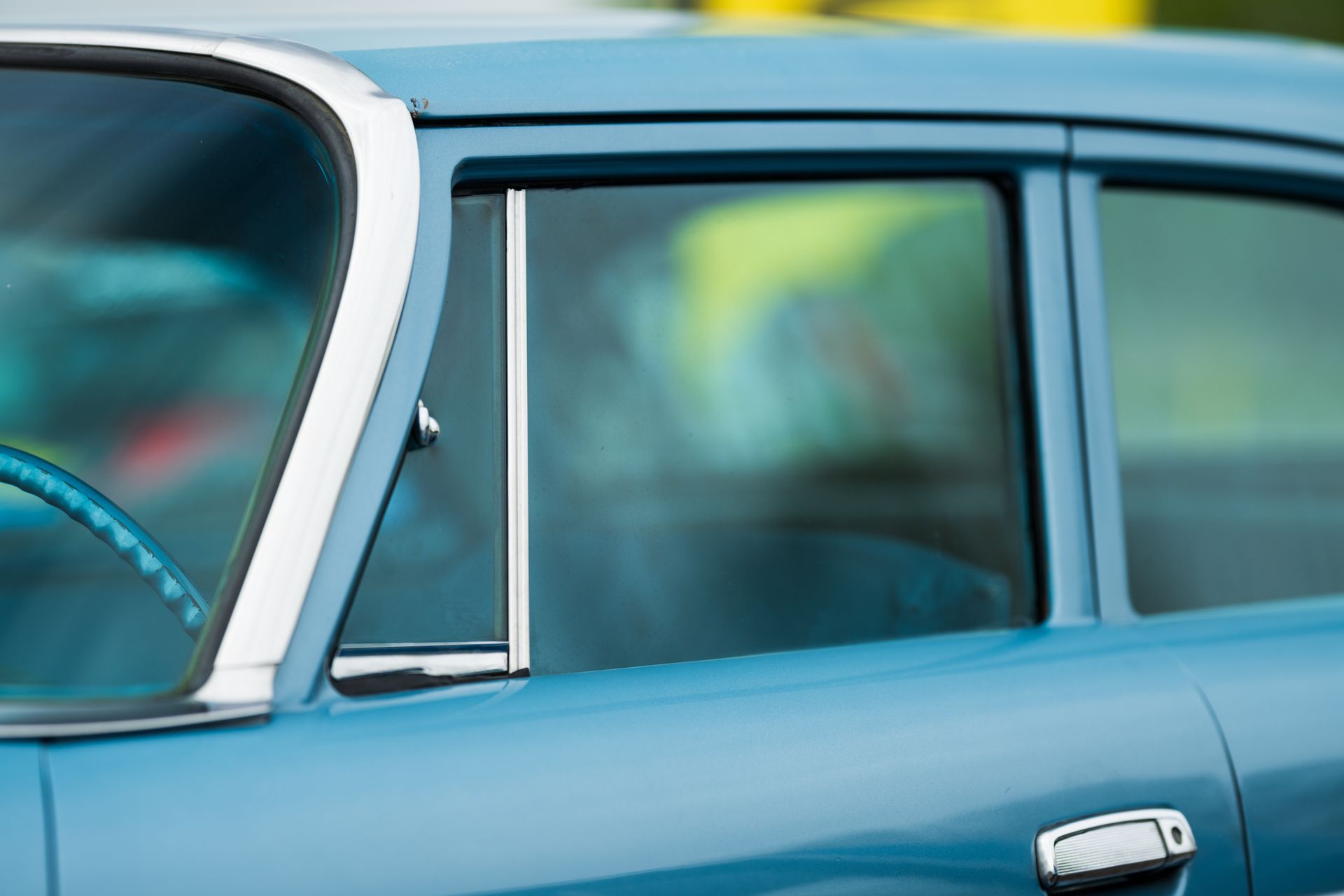 Close-up of a teal classic car window and door with silver trim; steering wheel visible.