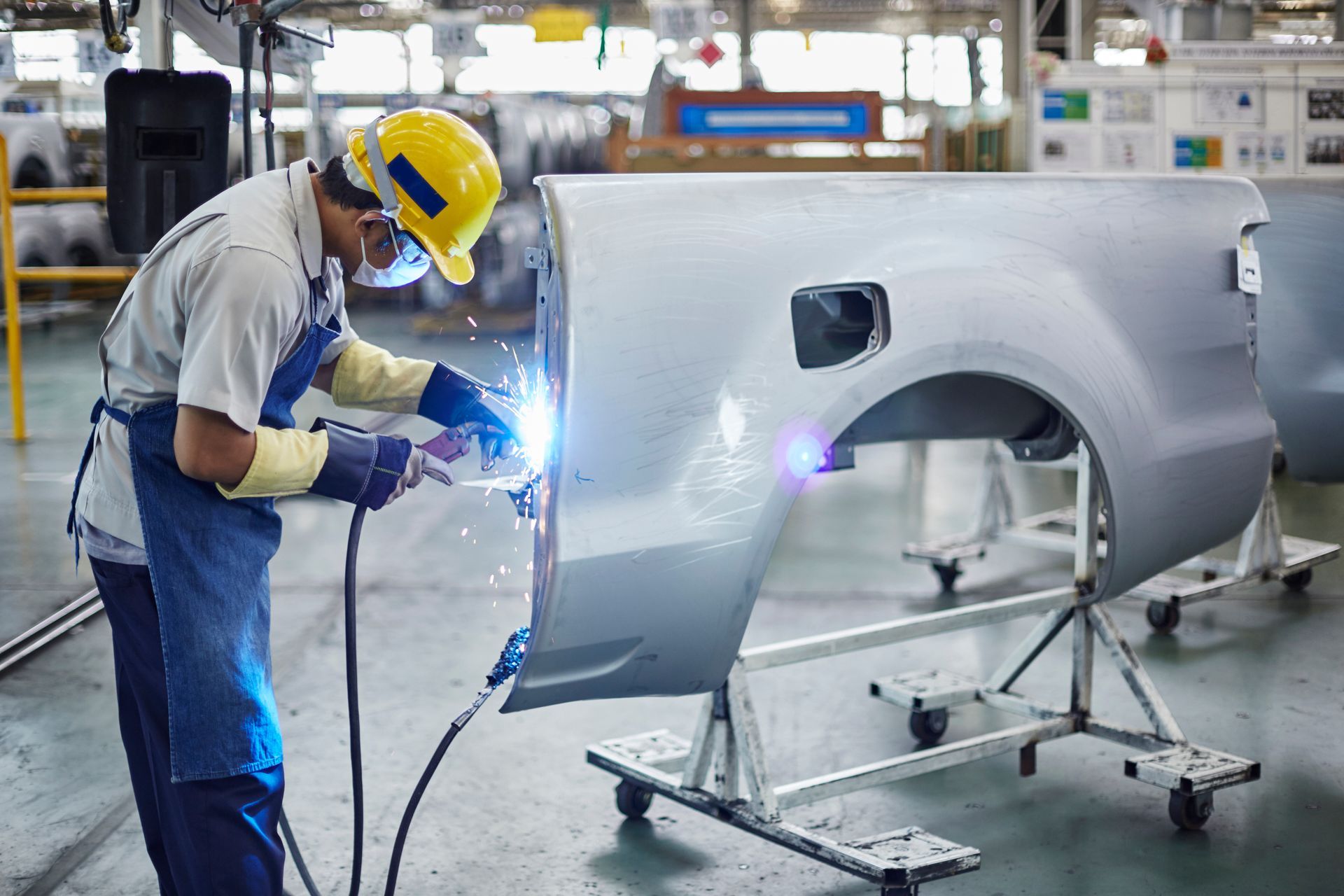 Welder in yellow hard hat welding car part in a factory setting.