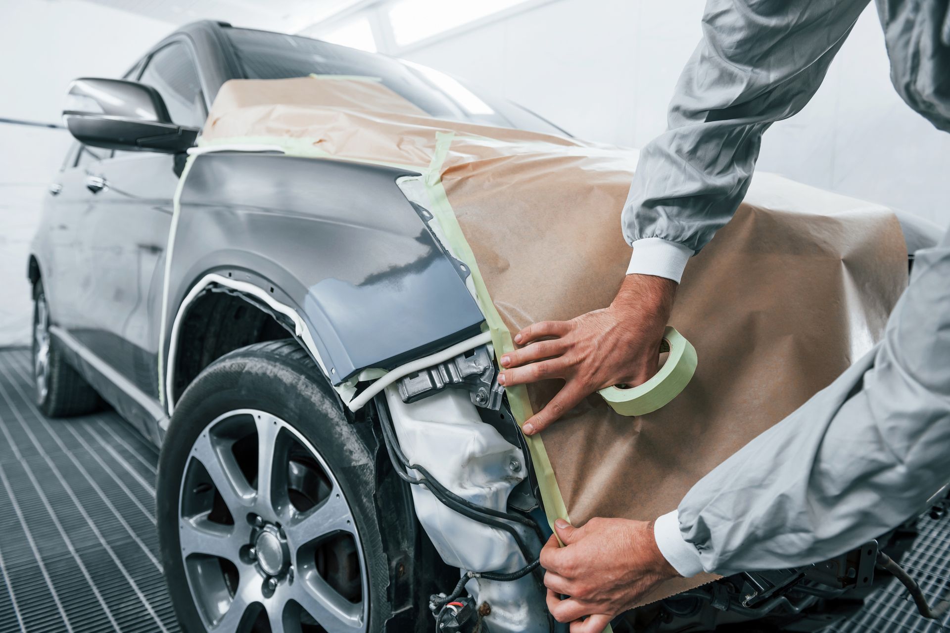 Person in a jumpsuit taping paper to a gray car's front fender, preparing for paintwork in a garage.