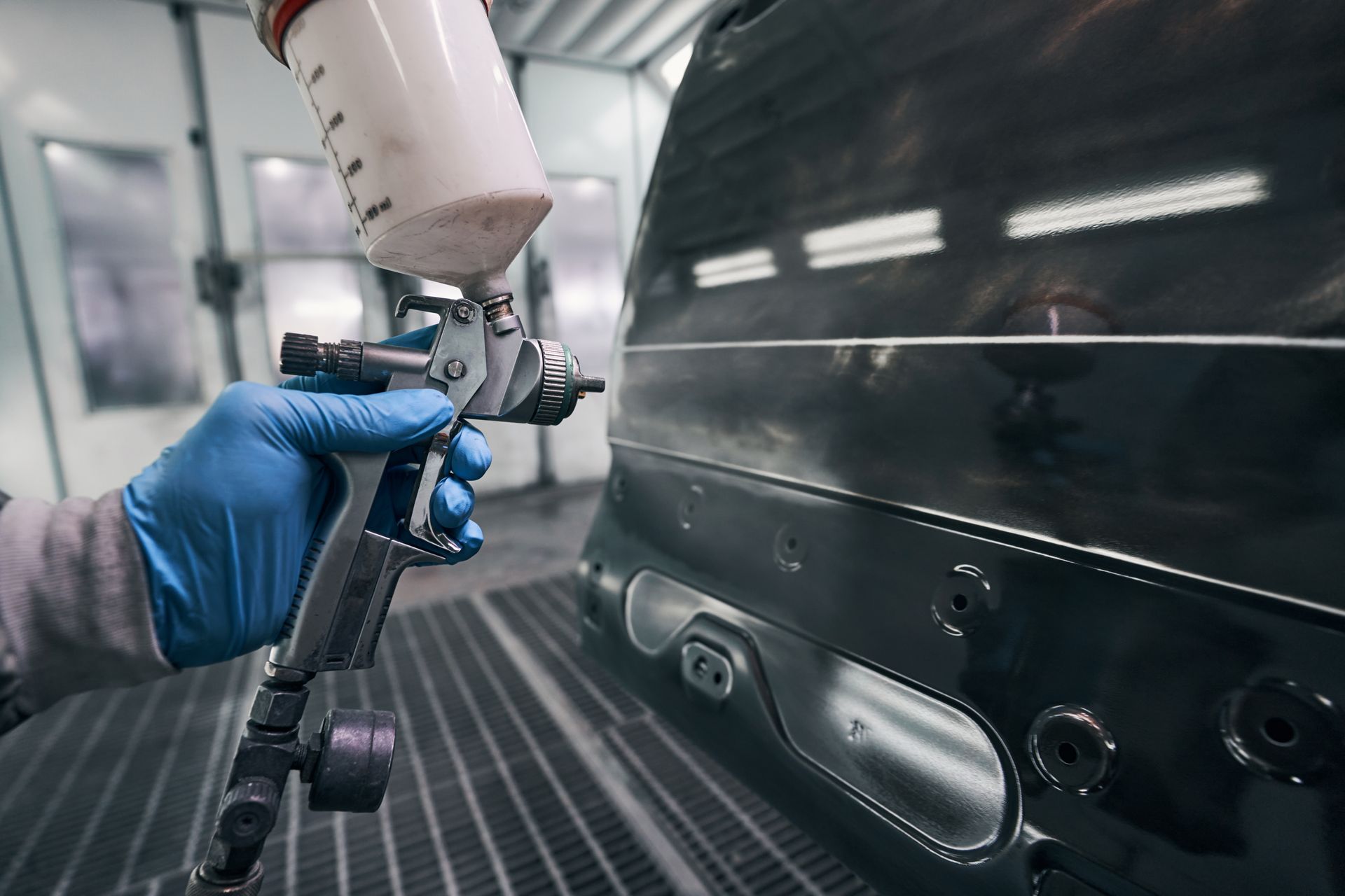 A person in blue gloves sprays paint on a car panel with a paint gun inside a spray booth.