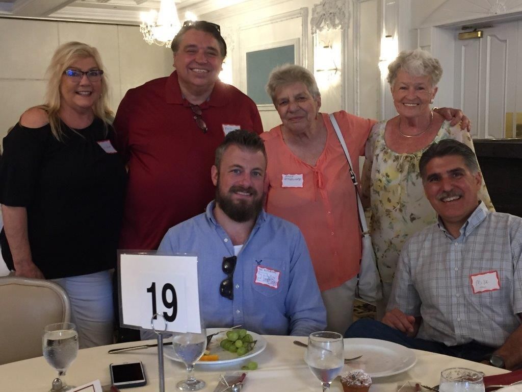 Group of six people posing at a dining table with a table number 19 in a bright room.
