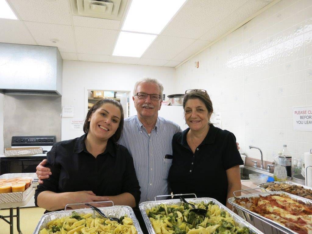Three smiling people behind a buffet table with trays of food in a bright kitchen