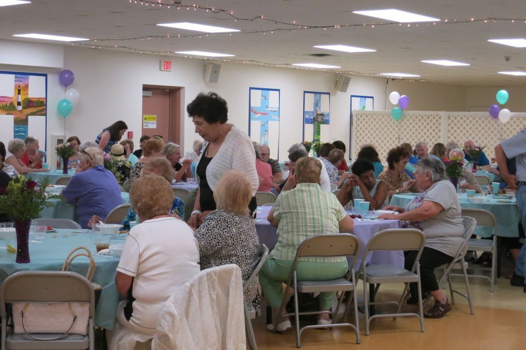 People seated at tables in a community hall, with balloons and a man standing among them.