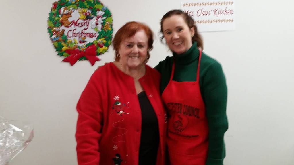 Two women in red aprons smiling at a decorated wall display in a bright room