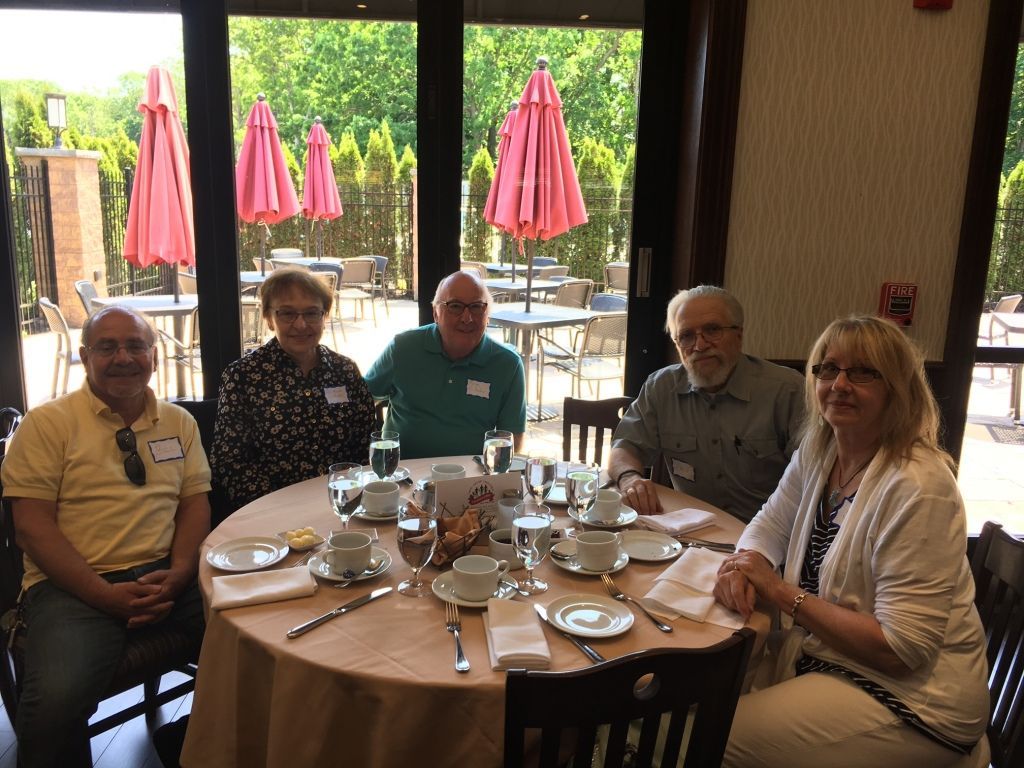 Five people seated around a restaurant table, smiling indoors with patio umbrellas visible outside.