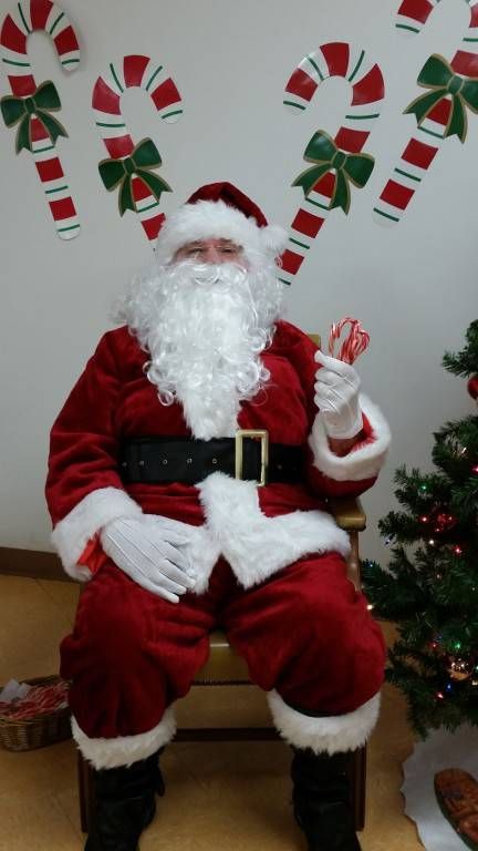 Santa in a red suit seated indoors with candy cane wall decor and a small Christmas tree nearby