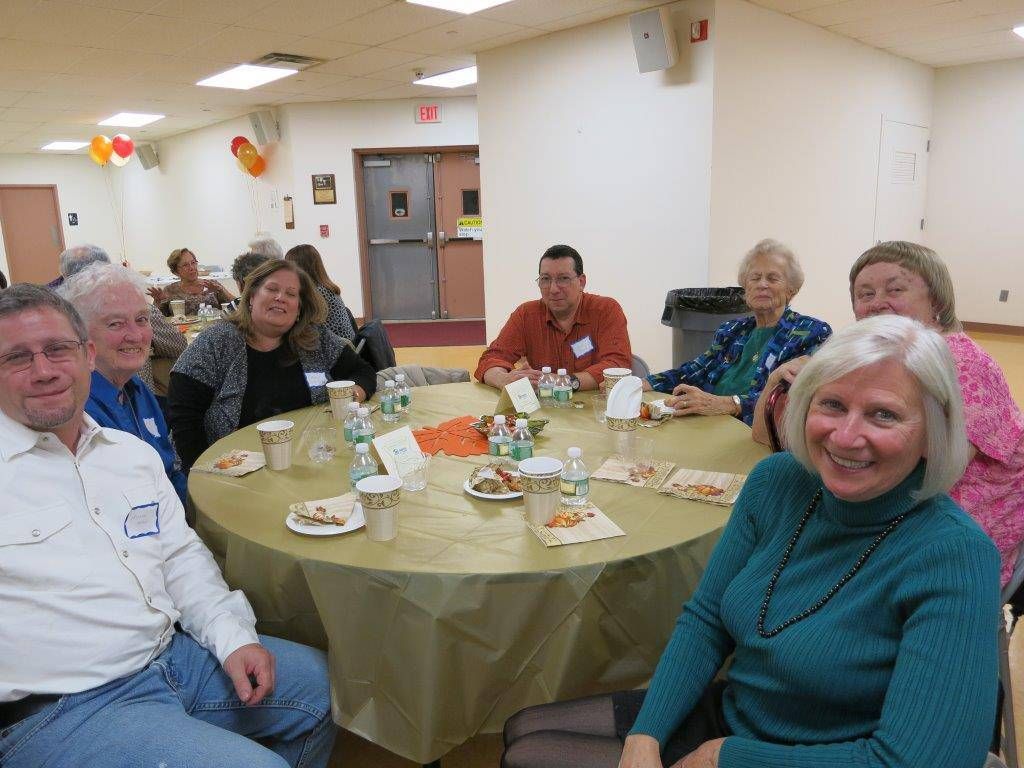 People seated around a round table in a community hall, chatting over drinks and snacks