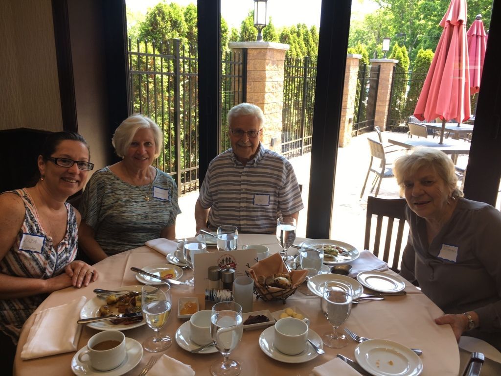 Four people seated around a table at an outdoor café, with cups, plates, and a garden view behind them.