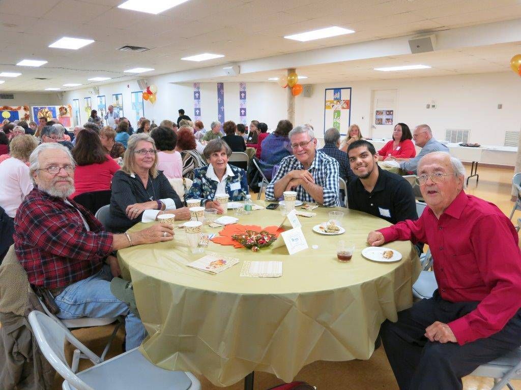 People seated at decorated banquet tables in a bright community hall, sharing a meal and conversation.