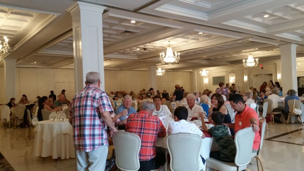 People seated at round tables in a banquet hall listening to a speaker in a plaid shirt.