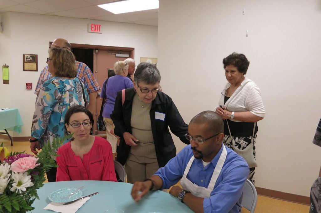 People seated at a table in a community room while others stand and talk nearby.