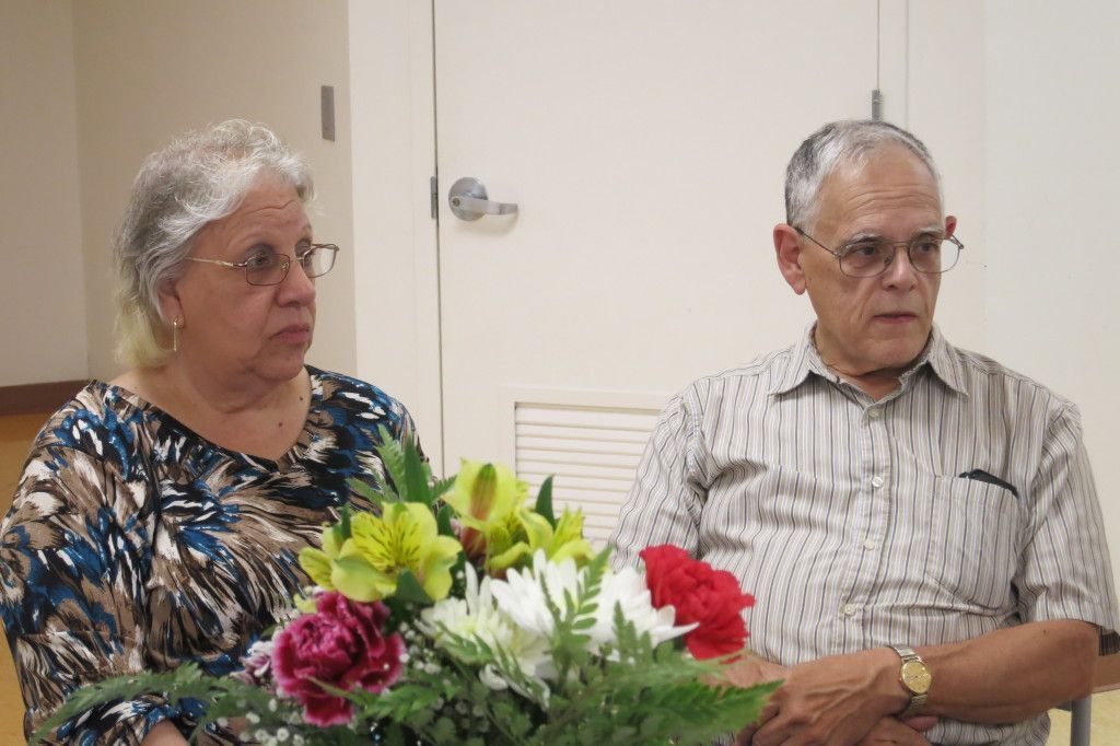 Two people seated indoors beside a colorful flower arrangement, looking off to the side.