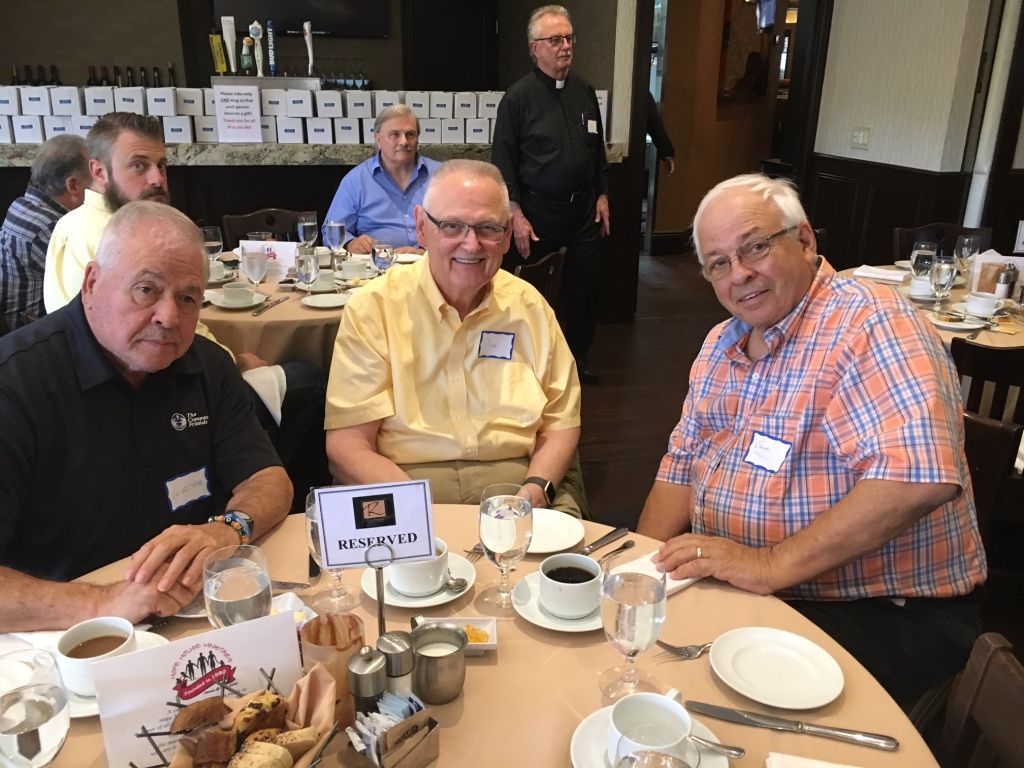 Three men seated at a dining table with coffee cups in a restaurant, smiling at the camera.