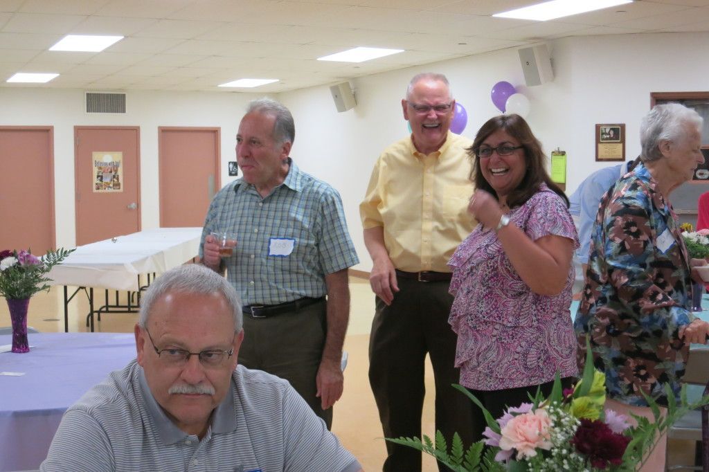 People standing and smiling at a community hall gathering, with flowers and tables in the foreground