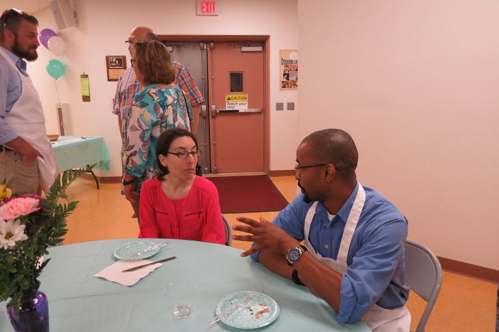 People talking at a round table in a community room, with flowers, papers, and a doorway in the background.