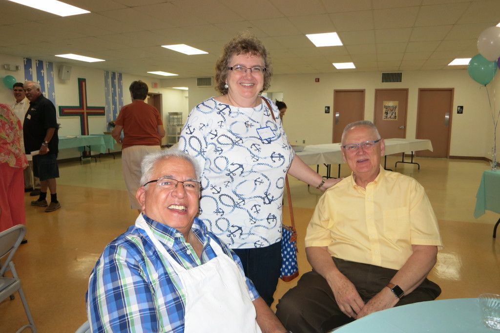 Three people smiling in a community room, with balloons and tables in the background.
