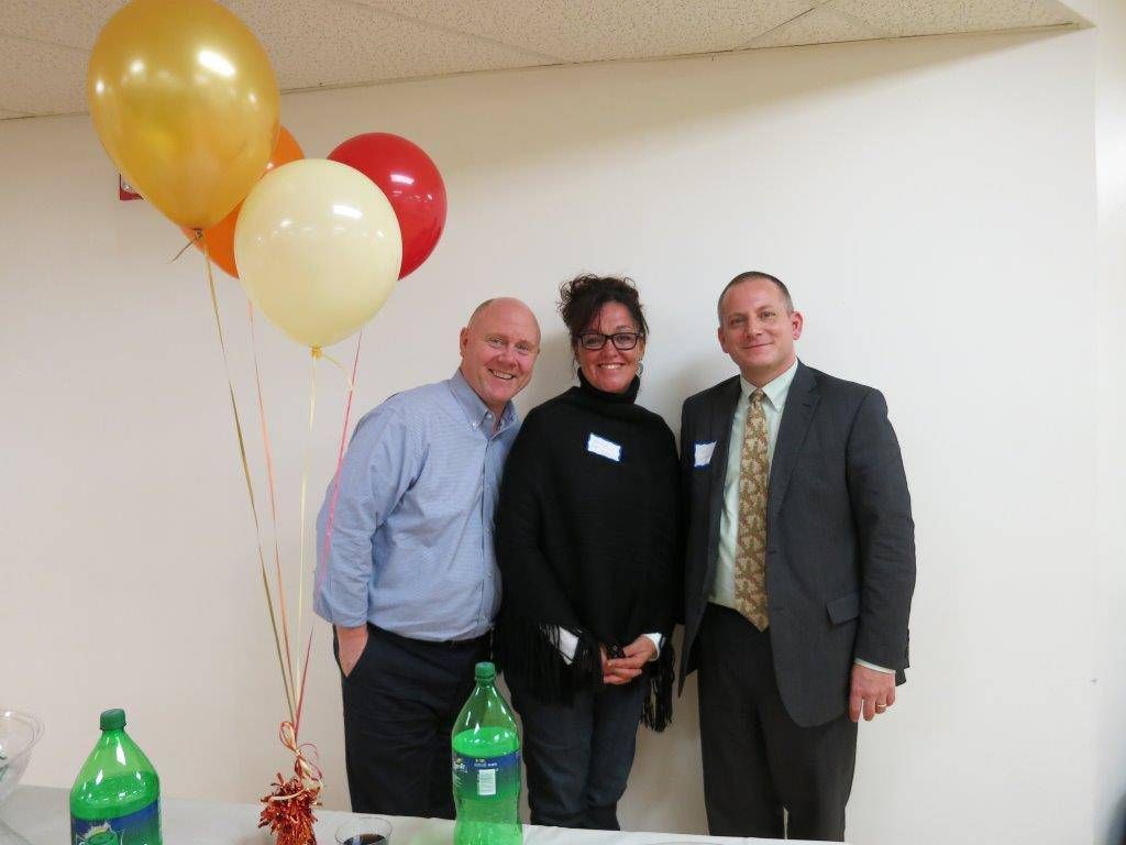 Three people posing indoors beside gold, white, and red balloons at a small celebration.