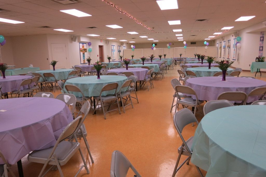 Large banquet hall with round tables, metal chairs, and pastel tablecloths under bright ceiling lights