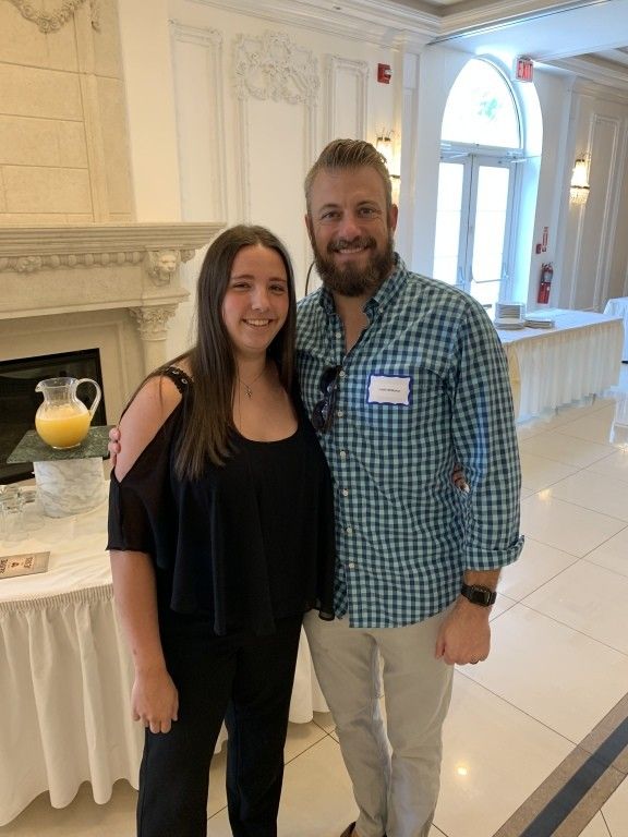 Two smiling people stand together in a bright indoor lobby beside a white fireplace.