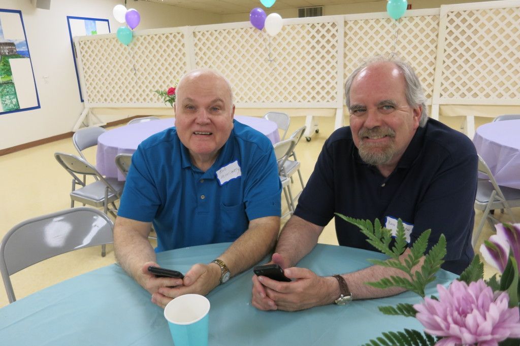 Two men seated at a decorated table, smiling and holding phones in a bright room with balloons.