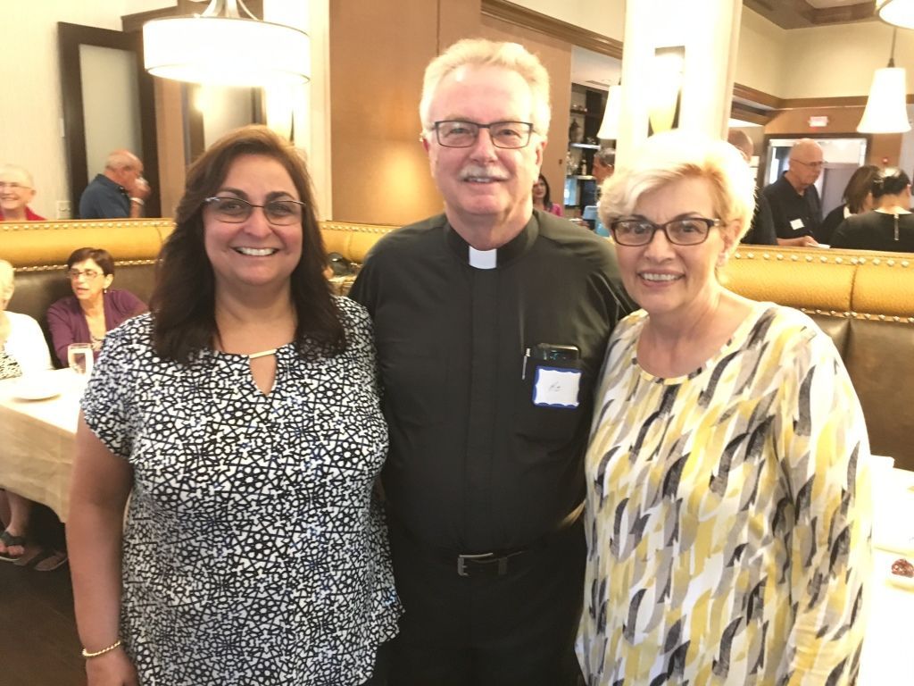 Three people posing indoors, with a priest in clerical attire centered between two smiling women.
