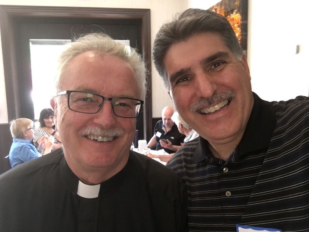 Two smiling men in a restaurant, one in a clerical collar and glasses, posing for a selfie.