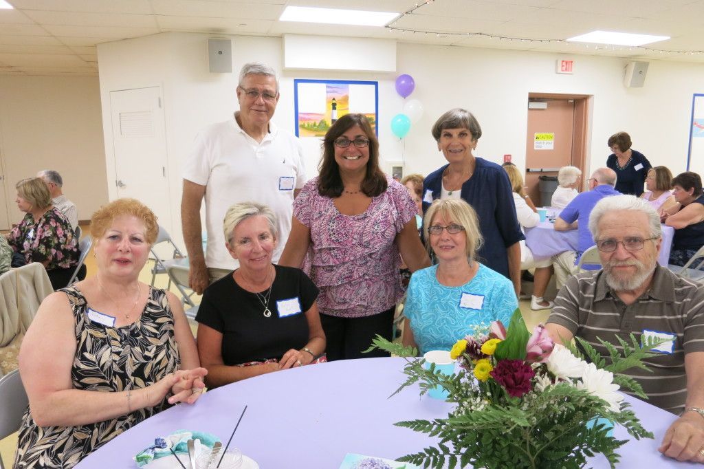 Group of people seated and standing around a table at an indoor gathering, with flowers in the center.