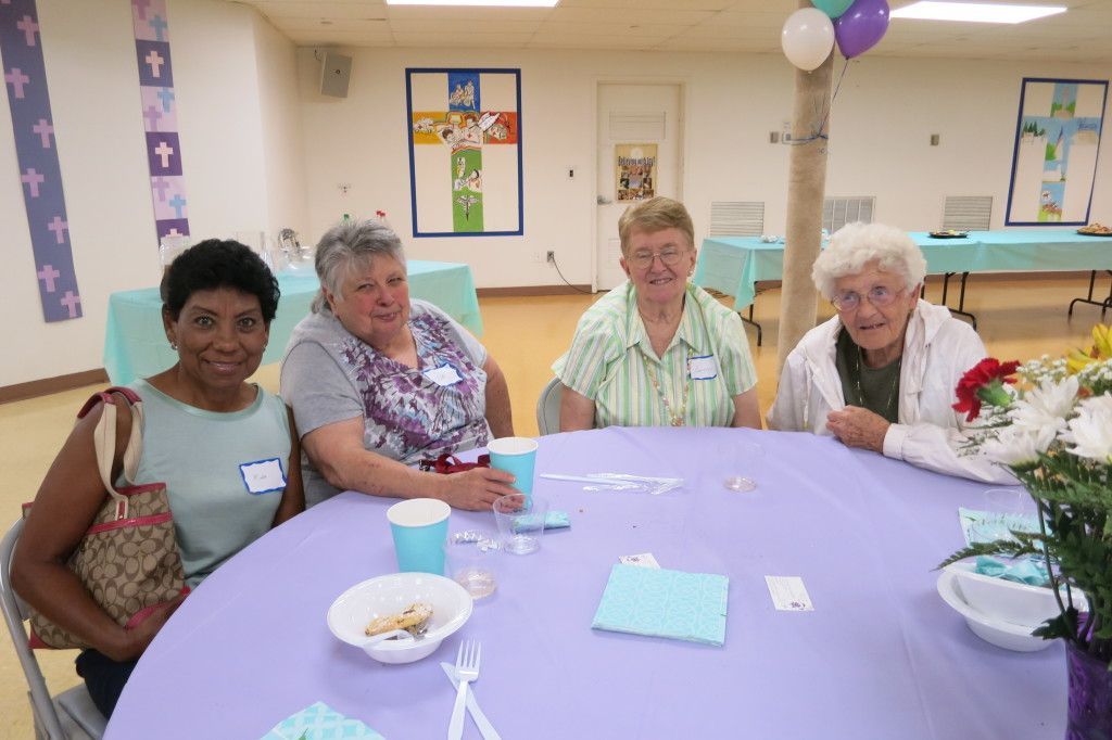 Four people seated at a purple table in a decorated community room, smiling at the camera.