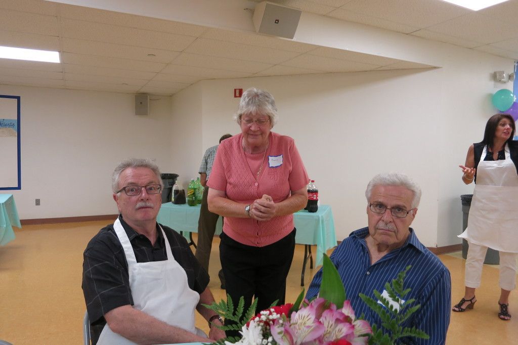 Two older adults seated at a table with flowers, with a woman standing behind them in a community room.