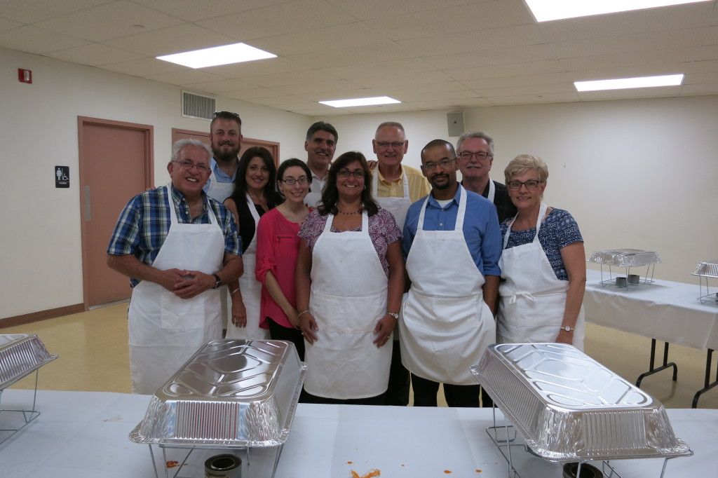Group of people in aprons posing behind food trays in a bright cafeteria kitchen