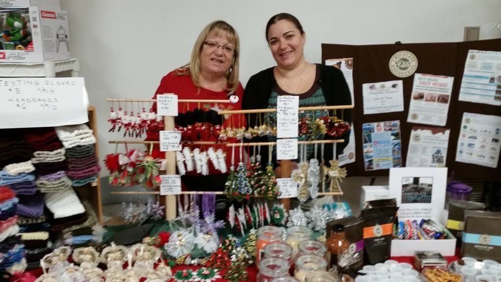Two vendors behind a craft fair table displaying handmade ornaments, jewelry, and gift items